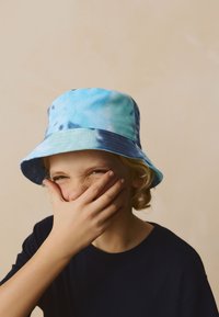 Vedierkový klobúk v modrom tie-dye vzore; mäkká tkanina so širokým okrajom; nosí ho dieťa, ktoré si rukou zakrýva ústa; vidieť tmavú košeľu.