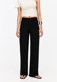 Pantalon noir à jambes droites avec une texture lisse, doté d'une fermeture à bouton et de passants de ceinture, associé avec un haut beige ajusté et des sandales.