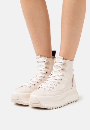 Pieds chaussés de baskets montantes crème à plateforme avec semelles épaisses à rainures et lacets blancs sur fond blanc.