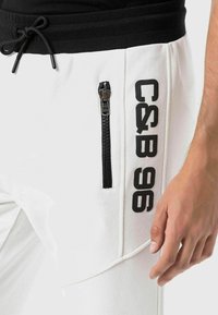 Weiße Jogginghose mit einem schwarzen elastischen Bund, versehen mit einer seitlichen Reißverschlusstasche und schwarzem Drucktext "C&B 96" entlang des Beins.