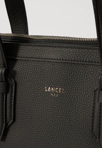 Black leather handbag with a textured finish, featuring a gold embossed logo "LANCEL PARIS" and a zip closure.