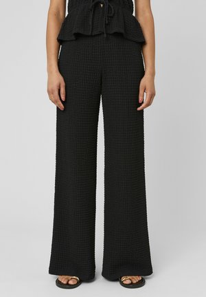 Femme portant un pantalon large noir texturé et un haut assorti, debout les mains détendues le long du corps, portant des sandales noires et dorées.