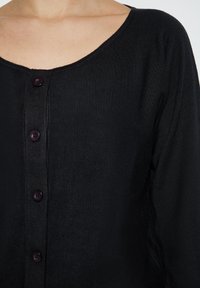 Černý kardigan s kulatým výstřihem a zapínáním na knoflíky. Má měkkou, strukturovanou tkaninu a pět tmavě fialových knoflíků jako dekoraci.