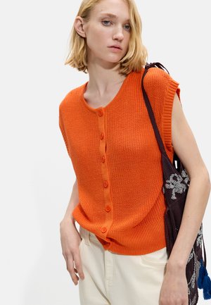 Blondynka w pomarańczowym, bezrękawnym, zapinanym na guziki, dzianinowym topie i jasnych spodniach, niosąca ciemną torebkę szydełkową z białymi wzorami i niebieskimi frędzlami.