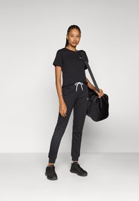 Black t-shirt with short sleeves, paired with black joggers featuring a white drawstring, and black athletic shoes. The model holds a black bag.