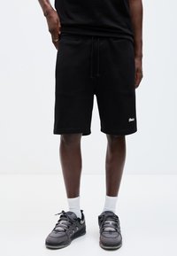 Modèle masculin portant un short noir à cordon, des chaussettes blanches et des baskets grises, debout devant un fond clair uni.