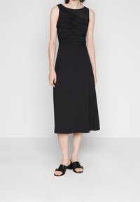 Femme portant une robe midi noire sans manches avec un corsage froncé et des sandales à talons ouverts noires, se tenant devant un fond blanc uni.