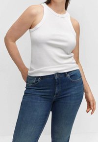 Woman wearing a sleeveless white ribbed top and high-waisted blue jeans, standing with one hand behind her back.