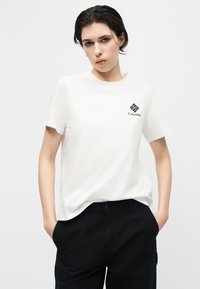 Νεαρός ενήλικας με κοντά σκουρόχρωμα μαλλιά, φοράει λευκό t-shirt Columbia και μαύρο παντελόνι, στέκεται με τα χέρια στις τσέπες σε απλό φόντο.