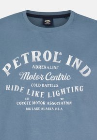 Modré bavlněné tričko s bílými grafickými nápisy. Nápisy zahrnují "PETROL IND," "Motor Centric" a "Coyote Motor Association." Klasický kulatý výstřih.