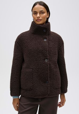 Frau trägt eine dunkelbraune, strukturierte Jacke mit Knöpfen, hohem Kragen und aufgesetzten Taschen, steht vor einem einfarbigen hellen Hintergrund.