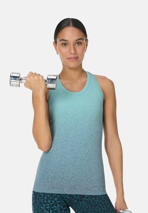 Vrouw in een blauwgroen sporttopje tilt met één arm zilverkleurige halters op, kijkt naar de camera, tegen een effen lichte achtergrond.