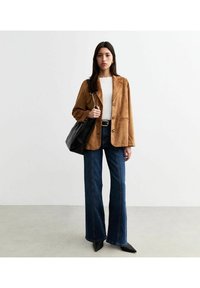 Bruine suède blazer over een witte top, gecombineerd met uitlopende blauwe jeans. Een grote zwarte tas met kettingdetails en puntige zwarte schoenen.