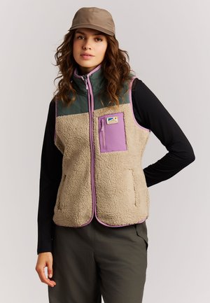 Frau trägt beige-lila Fleeceweste über schwarzem Langarmshirt und olivgrüner Hose mit beiger Kappe, steht mit einer Hand in der Hüfte.