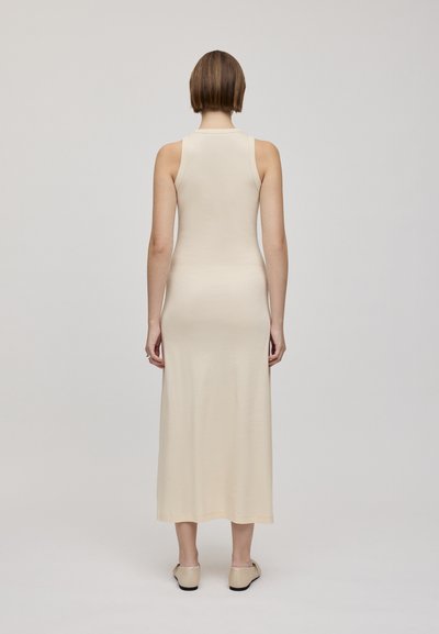 Abito beige senza maniche di lunghezza media realizzato in tessuto liscio, caratterizzato da un collo alto e una silhouette aderente, abbinato a calzature chiare.