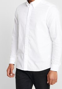 Hombre vestido con una camisa blanca de botones de manga larga y pantalones negros, de pie contra un fondo claro liso.