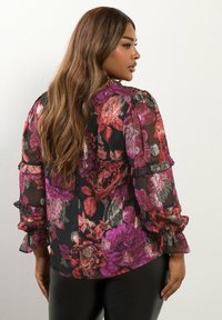 Blusa nera trasparente con motivo floreale in viola, rosso e accenti metallici. Maniche a sbuffo e dettagli sulla schiena con una vestibilità comoda.