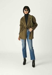 Vrouw in een olijfgroene belted coat, zwarte coltrui, blauwe jeans, zwarte enkellaarsjes, zonnebril en met een groene schoudertas.