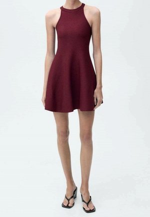 Kvinde iført ærmeløs burgundy halterneck kjole og sorte kilehælede sandaler, stående mod en ensfarvet lys baggrund.