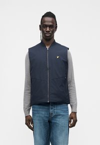 Tengerészkék mellény, elöl cipzárral, bal mellrészen arany logóval. Világosszürke hosszú ujjú póló és kék farmer nadrág fölött viselve.