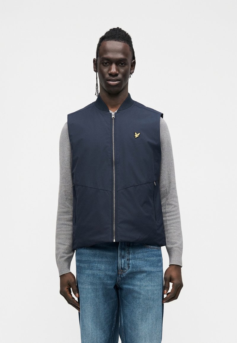 Marineblauw vest met een ritssluiting aan de voorkant, met een gouden logo op de linkerborst. Draagt over een lichtgrijs shirt met lange mouwen en blauwe spijkerbroek.