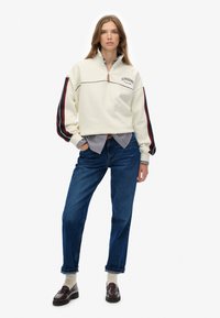 Crème kleurige sweatshirt met rits en zwart-rode gestreepte mouwen, gecombineerd met blauwe straight-leg jeans en donkere loafers over crème sokken.