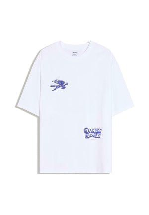 Fehér rövid ujjú póló kék madár grafikával a bal mellrészen és kék szöveggel, amely így szól: "SAJÁT ÚTAMON HALADOK" a jobb alsó részen.