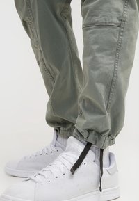 Gröna cargobyxor med elastiska knytningar och sidofickor, parat med vita sneakers med strukturerad tyg och svarta snören som accent.