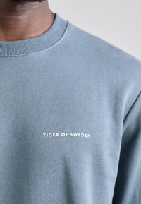 Primer plano de una persona que lleva un suéter azul claro de cuello redondo con "Tiger of Sweden" impreso en blanco en el pecho.
