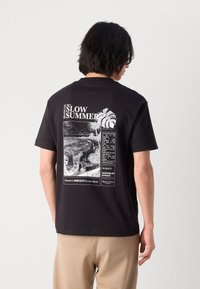 Black cotton t-shirt featuring a graphic print on the back with tropical imagery and text about a "Slow Summer." Short sleeves, crew neck design.