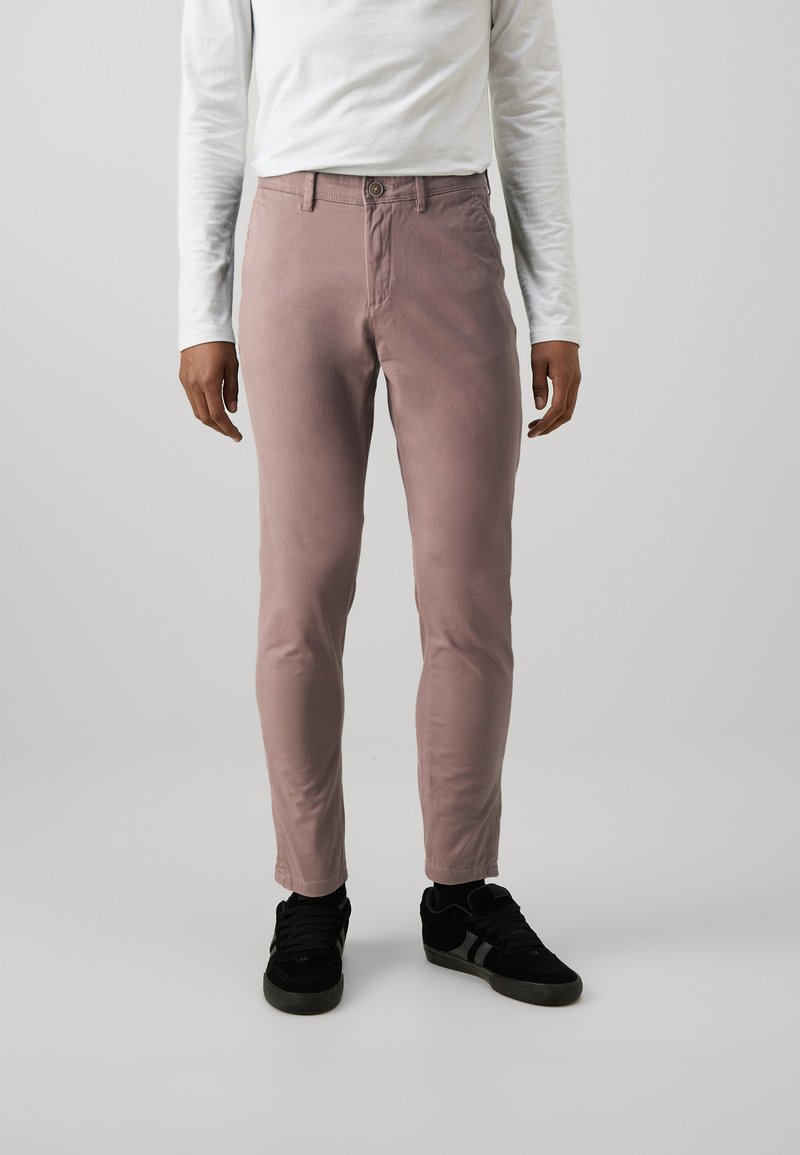 Pantalones de algodón rosa claro con un ajuste ceñido, que cuentan con un cierre de botón y bolsillos, combinados con zapatillas negras. Textura suave, diseño minimalista.