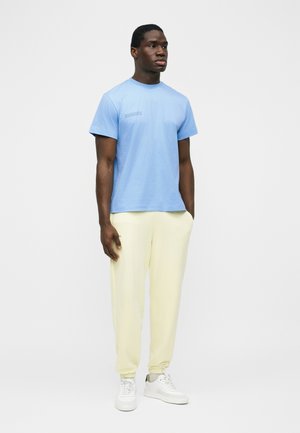 Mann steht mit den Händen in den Taschen einer hellgelben Jogginghose, trägt ein hellblaues T-Shirt mit kurzen Ärmeln und weiße Sneakers vor einem schlichten Hintergrund.