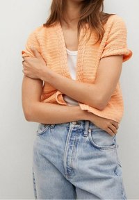 Oranger Strickcardigan mit kurzen Ärmeln, strukturiertem Muster, offener Front, über einem weißen Tanktop getragen, kombiniert mit hochgeschnittenen hellblauen Jeans.