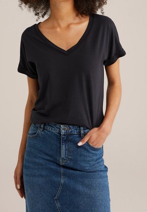 Kvinde iført en sort V-hals kortærmet T-shirt, stoppet ned i et blåt denimskørt med den ene hånd i skørtets lomme.