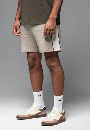 Beige Sportshorts mit schwarzem Seitenstreifen, kombiniert mit weißen Knöchelsocken, die ein schwarzes Logo aufweisen, und creme-braunen Sneakers.