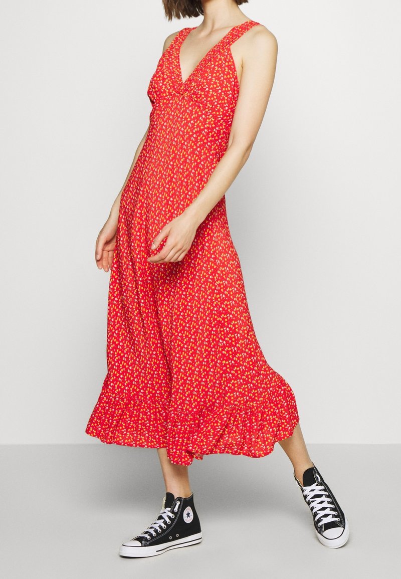Femme portant une robe midi sans manches à fleurs rouges, associée à des baskets montantes noires, debout devant un fond blanc uni.