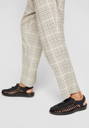 Personne portant un pantalon à carreaux beige et des sandales noires tissées, debout sur une surface gris clair.
