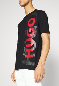 Sort t-shirt med et stort rødt "HUGO" grafisk design på venstre side. Lavet af bomuld med en afslappet pasform og rund udskæring.