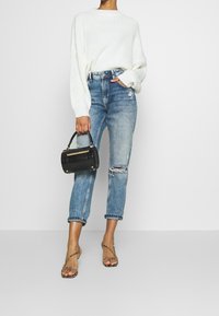 Femme portant un pull en maille blanc et un jean bleu déchiré, tenant un sac à main noir, portant des sandales à talons imprimé serpent, debout sur un sol gris.