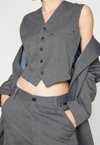 Szary w kratę krótki kamizelka z głębokim dekoltem w kształcie litery V, sześcioma czarnymi guzikami i kieszenią na piersi. Połączona z dopasowanymi szarymi spodniami o wysokim stanie.
