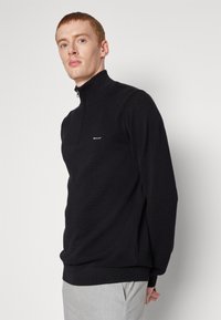 Zwarte half-zip pullover van textiel, met een klein logo op de borst en geribbelde manchetten en zoom, gecombineerd met lichtgrijze broeken.
