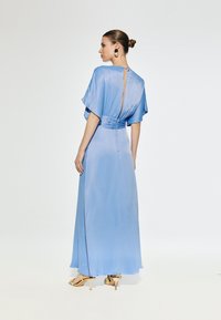 Duga, lepršava plava haljina od sjajnog materijala, s kratkim lepršavim rukavima, prorezom na leđima i uskim strukom. Zlatne sandale s remenčićima.