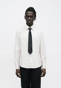 Muž s tmavou pokožkou a dredmi, oblečený v svetlej pruhovanej košeli, čiernej kravate a čiernych nohaviciach, stojaci pred jednoduchým bielym pozadím.