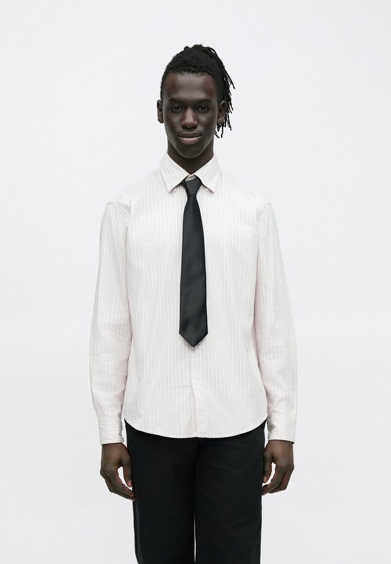 Muž s tmavou pokožkou a dredmi, oblečený v svetlej pruhovanej košeli, čiernej kravate a čiernych nohaviciach, stojaci pred jednoduchým bielym pozadím.