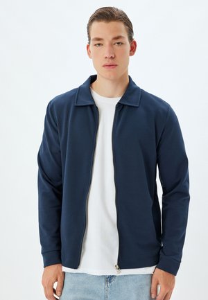 Marine blauwe zip-up jack met een kraag, lange mouwen en een lichtgewicht stof. Gekocht over een wit T-shirt, gecombineerd met blauwe denim jeans.