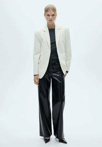 Witte op maat gemaakte blazer over een donkere top, gecombineerd met glanzende zwarte brede broek met een krokodillenstructuur en puntige zwarte hakken.
