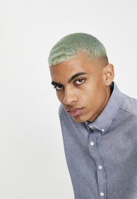 Jeune homme aux cheveux courts vert clair portant une chemise bleue claire boutonnée, regardant intensément la caméra sur un fond blanc uni.