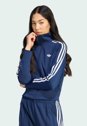Kobieta ubrana w granatowy dres Adidas z białymi paskami, zapiętą na całej długości do szyi, pozująca z dłonią blisko twarzy i poważnym wyrazem.