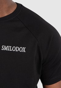 Czarny bawełniany t-shirt z okrągłym dekoltem i krótkimi rękawami raglanowymi. Posiada białe haftowane logo "SMILODOX" na klatce piersiowej.