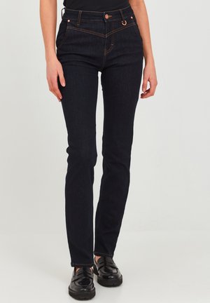 Jean en denim bleu foncé avec une coupe slim, ornés de couture orange, de poches avant et d'une fermeture à bouton. Portés avec des chaussures noires.